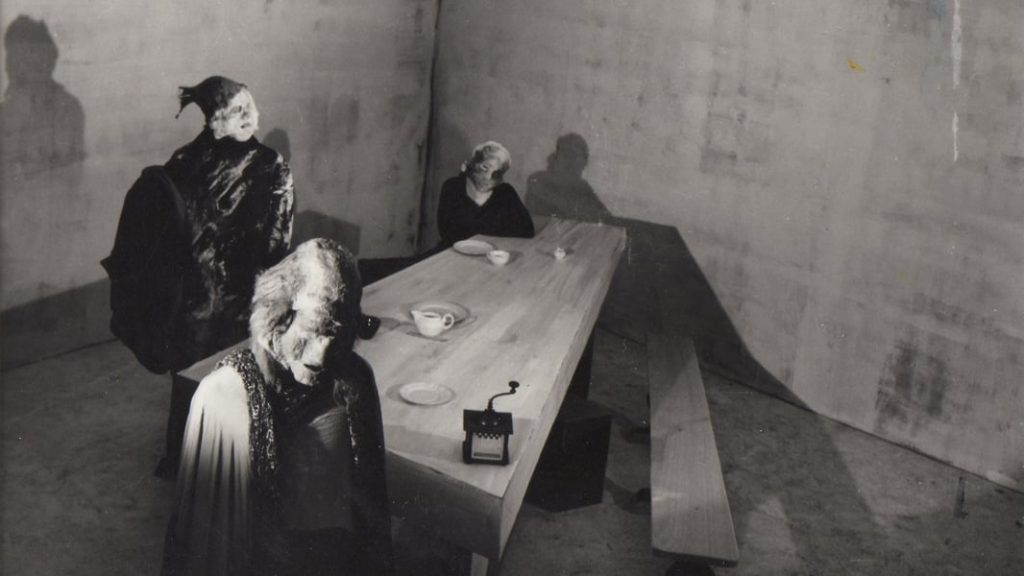 Photogramme issu de LA FEMME QUI SE POUDRE : trois personnages masqués et informes se tiennent autour d'une longue table en bois 