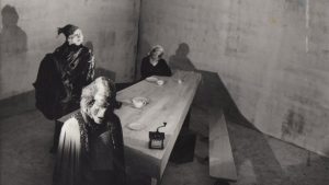 Photogramme issu de LA FEMME QUI SE POUDRE : trois personnages masqués et informes se tiennent autour d'une longue table en bois