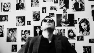 Jean-Luc Godard, regard vers le haut devant un mur couvert de photos de tournage.