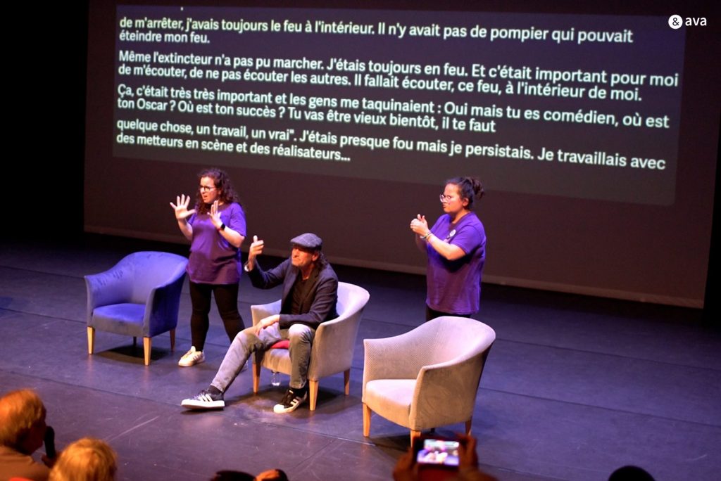 photographie d'une conférence avec deux interprètes LSF avec un sous titrages en direct sur un grand écran