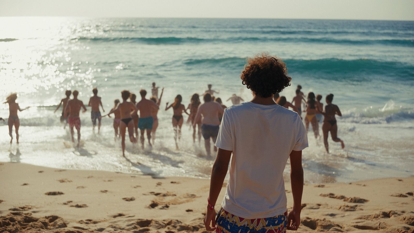 Un homme regarde de dos la plage et la mer, des gens courent vers l'eau